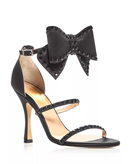 A black satin sandal featuring a deconstructed bow detail on the ankle strap, with a high heel and round toe.